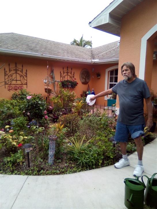 A man is watering colorful plants in a lush garden surrounded by cheerful blooms.
