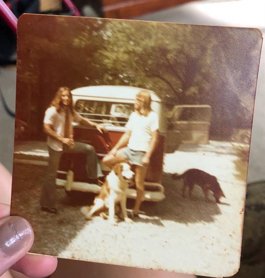 Two friends stand beside a vintage car in a rustic outdoor area, surrounded by dogs.