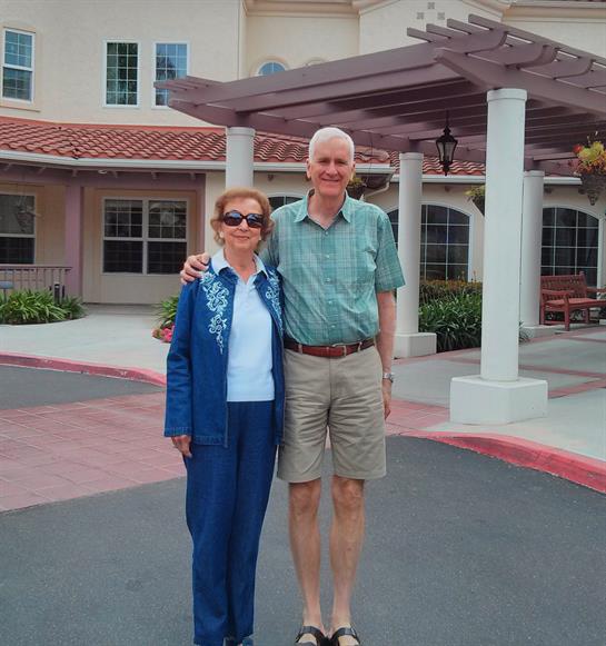 A smiling couple stands together on a pathway outside a senior living facility, enjoying their day.