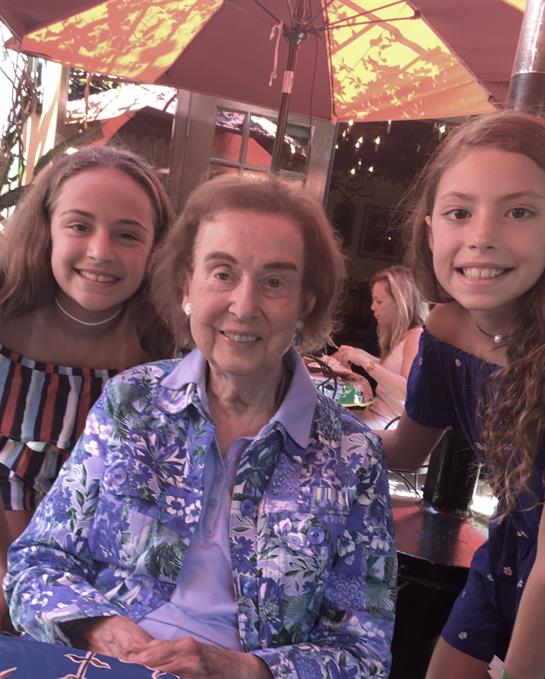 Two girls smile widely alongside a senior woman, enjoying a cheerful moment at an outdoor cafe.