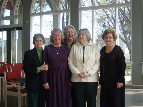Five women stand together, smiling and dressed in stylish attire, in a bright indoor setting.