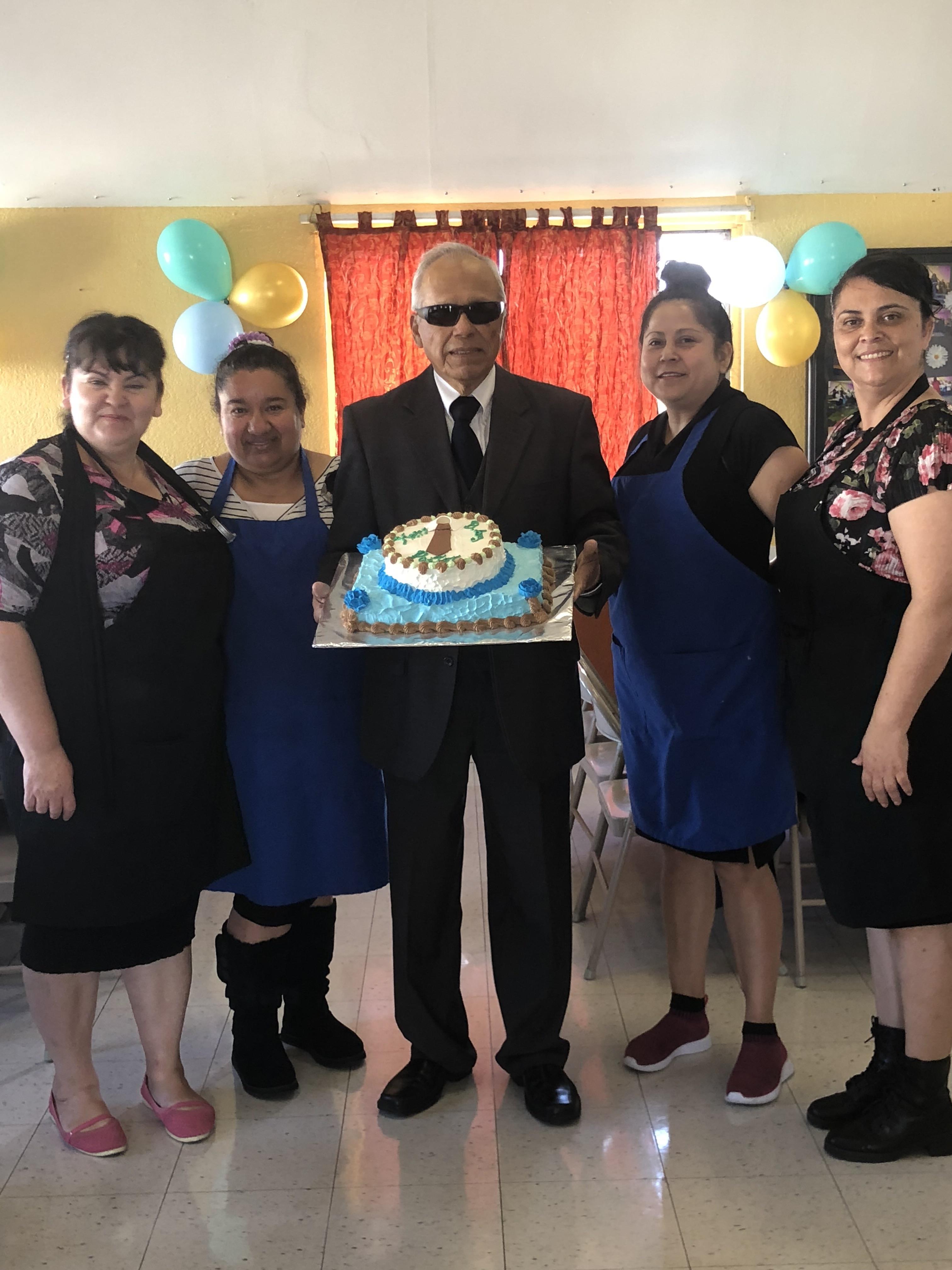 Family and friends gather for a joyful celebration, presenting a cake to a distinguished guest.