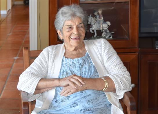 A senior woman sits comfortably in a chair, smiling and relaxed in her cozy living space.