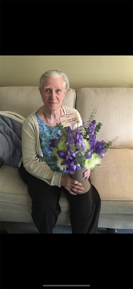 An elderly woman smiles while holding a colorful bouquet of flowers on her couch at home.