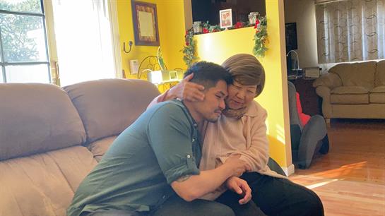 A young man and an older woman share a warm embrace on a couch in a cheerful living room.