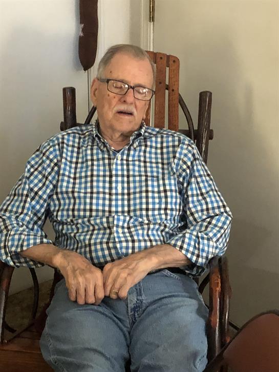 An elder man in a checkered shirt sits in a rocking chair, appearing relaxed and contemplative.