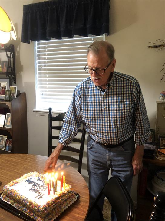 Older man lights candles on a birthday cake, surrounded by family photos and a warm atmosphere.