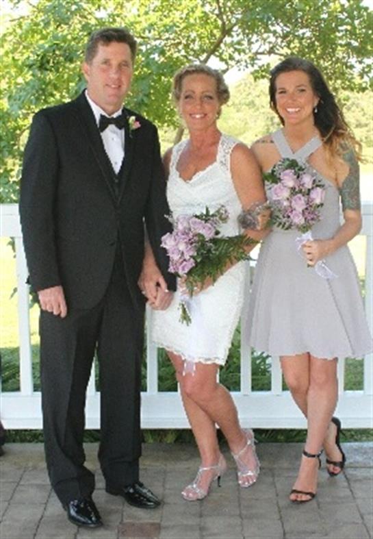 A couple in formal attire stands with a bridesmaid, all smiling and holding floral bouquets.