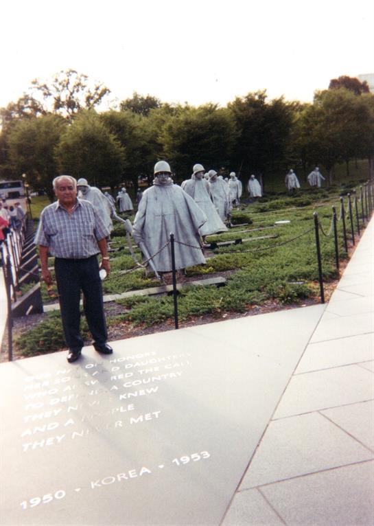 An elderly man stands by a unique memorial of military figures, set amidst greenery.