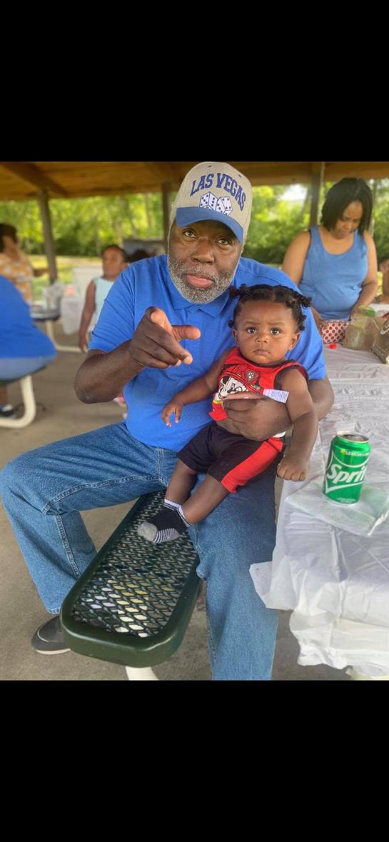 An elderly man in a blue shirt holds a baby at a picnic, surrounded by family and friends.