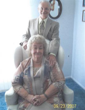 A man in formal wear stands behind a smiling older woman in a cozy chair, sharing warmth.