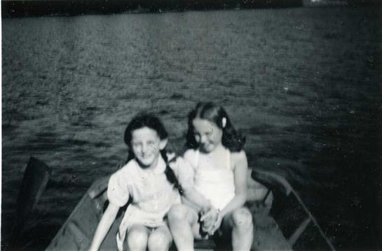 Two young girls sit side by side in a small boat on a serene lake, smiling and enjoying the day.