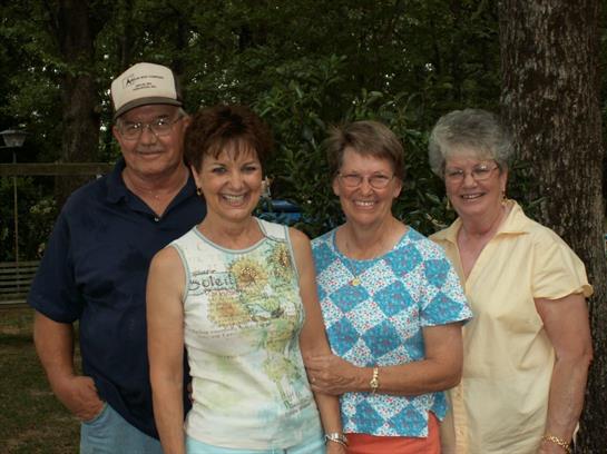 Four friends stand closely together, smiling and enjoying a warm day outdoors surrounded by nature.