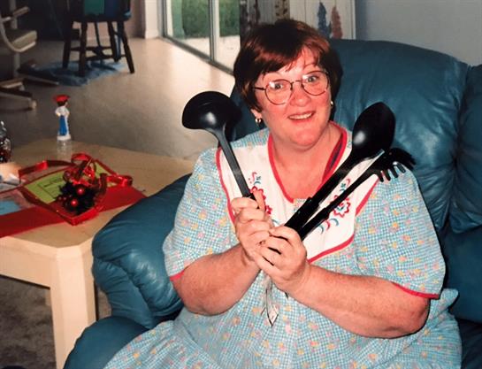 A woman in a blue dress smiles playfully while holding large kitchen utensils in a sunny room.