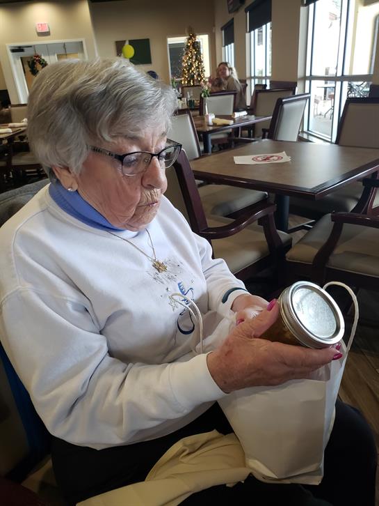 An older woman sits in a bright restaurant, focused on a jar in her hands, reflecting thoughtfully.