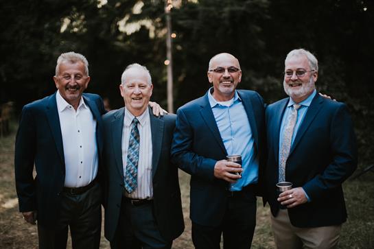 Four men in suits smile and hold drinks on a friendly summer evening.