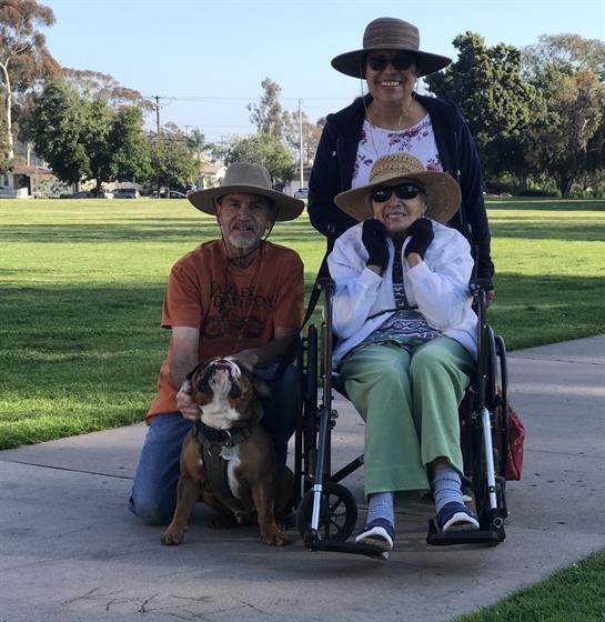 A family gathers in a park on a sunny day, smiling while interacting with a dog and a wheelchair.
