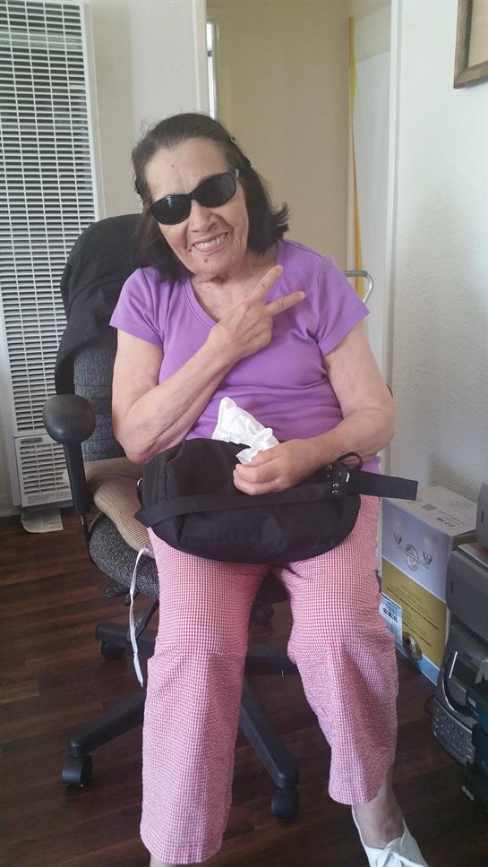 An elderly woman in sunglasses and a purple shirt poses playfully in her living room.