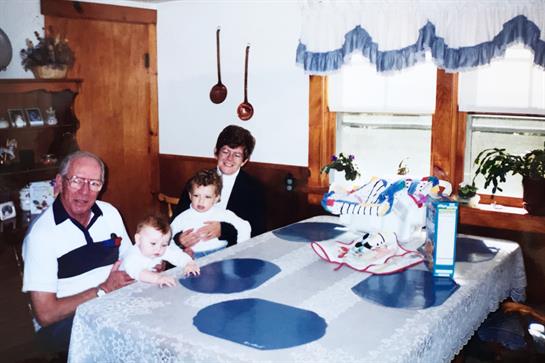 Two children are seated at a blue and white table, while an adult holds one and smiles.
