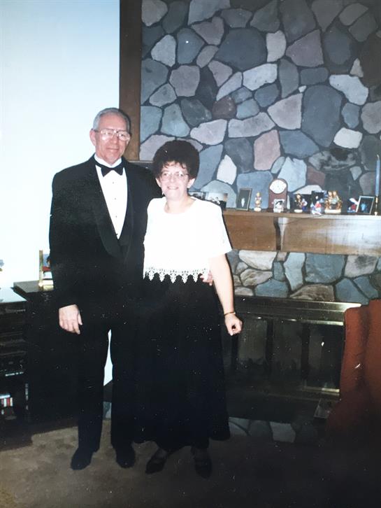 A couple in formal wear smiles happily together by a stone wall in their cozy home.