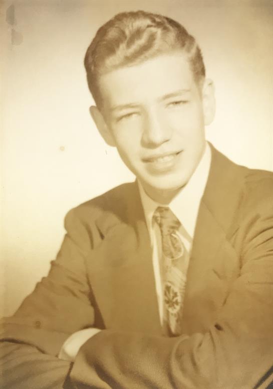 A young man sits confidently with a smile, wearing a suit and tie typical of the early 1960s.