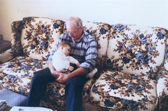A grandfather nestles a sleeping child in his arms while sitting on a floral couch at home.