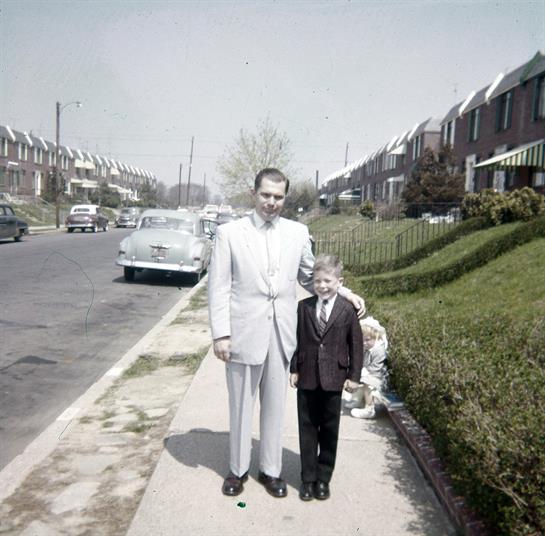 A man in a light suit and a boy in a dark suit pose on the sidewalk in a suburban neighborhood.