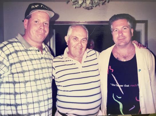 Three men stand close together smiling in a relaxed indoor social gathering.