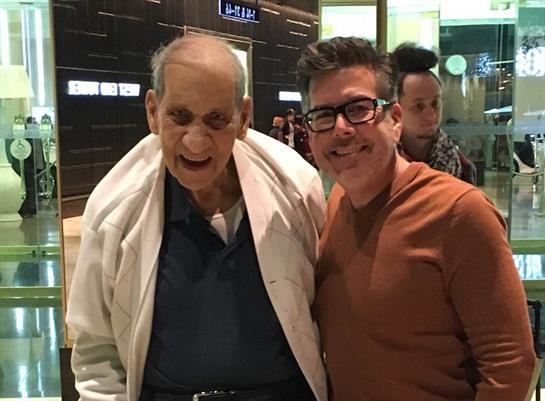 Two men smile while standing close together in a busy public area, enjoying their time.