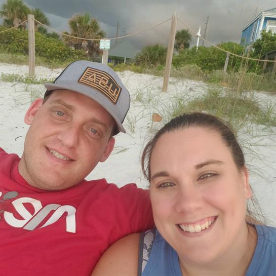 A couple sits comfortably on the sandy beach, smiling together as clouds gather overhead.