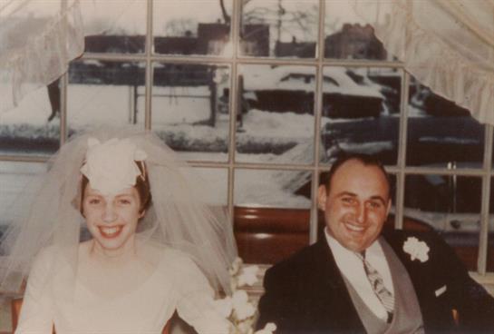 Couple joyfully poses on their wedding day with snow visible outside a large window.