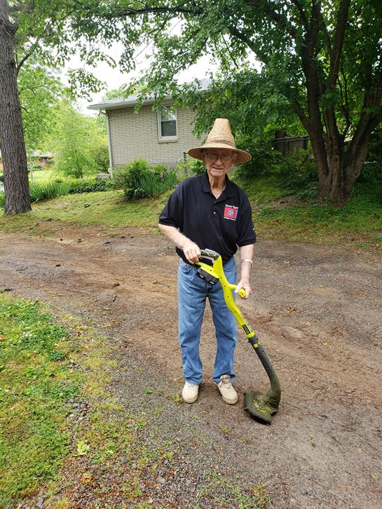 An elderly man in a hat operates a grass trimmer while maintaining his yard.
