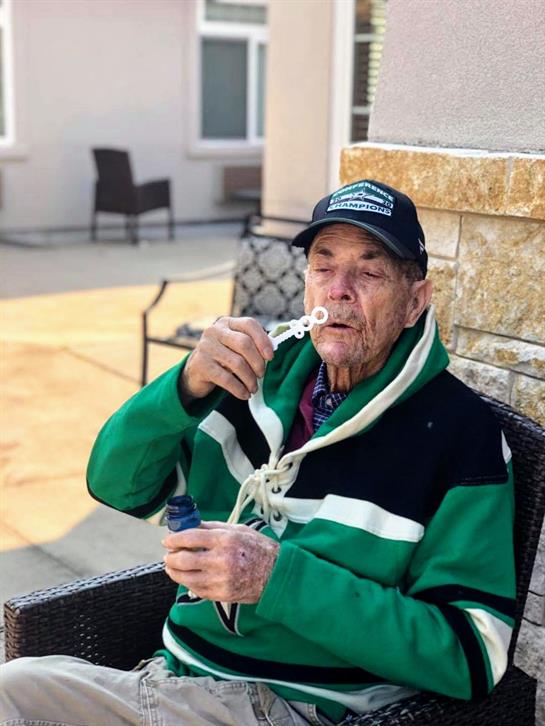 Man in a green jacket blows bubbles in a sunny courtyard, capturing a joyful moment outdoors.