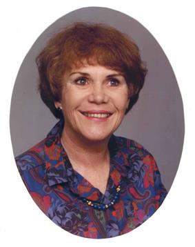 A woman with short hair smiles warmly while wearing a vibrant, patterned blouse.