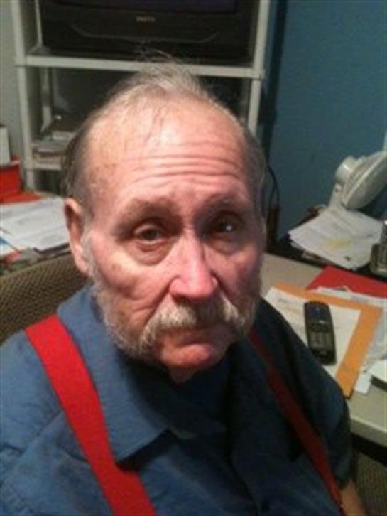 Old man wearing red suspenders looks surprised while sitting at a busy home office desk.