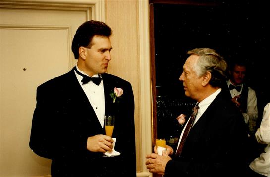 Two well-dressed men share drinks and converse at a high-end gathering in the evening.