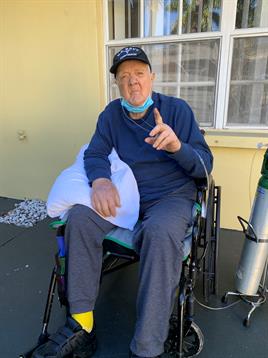An elderly man in a wheelchair smiles and gestures with one finger outdoors, enjoying the sunlight.