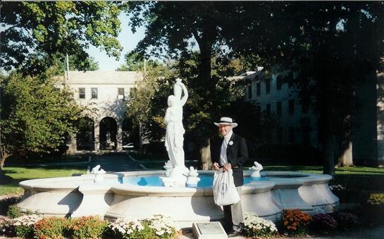 A man dressed in formal attire stands next to a statue fountain surrounded by lush greenery.