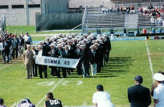 Graduates in uniforms parade across the field carrying a banner during their ceremony.