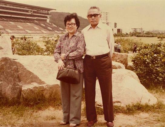 Elderly couple stands together on a rock, smiling and enjoying a sunny day in a park setting.