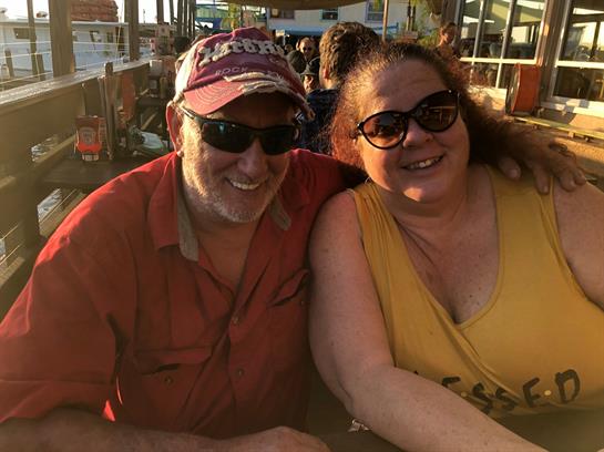 A smiling couple sits close, enjoying a warm sunset at a festive outdoor gathering.