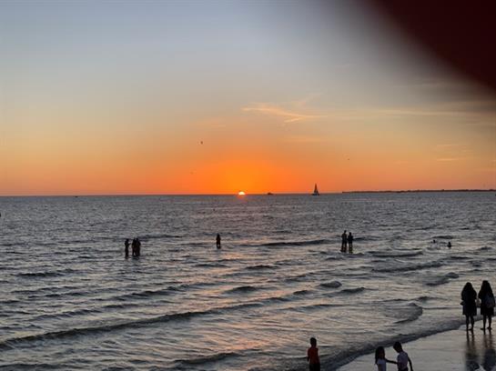 People enjoy the shoreline while watching the sun set over the calm ocean waters.