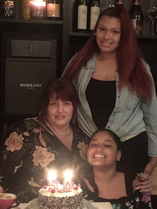 Three generations of women gather around a decorated cake with candles in a cozy room.