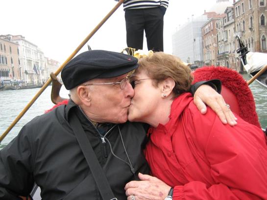 An elderly couple shares a tender kiss on a gondola, surrounded by a serene cityscape and mist.