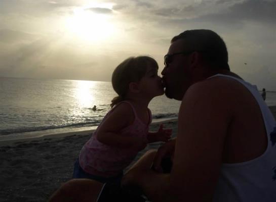 A girl leans in to kiss her father as the sun sets over the ocean horizon.