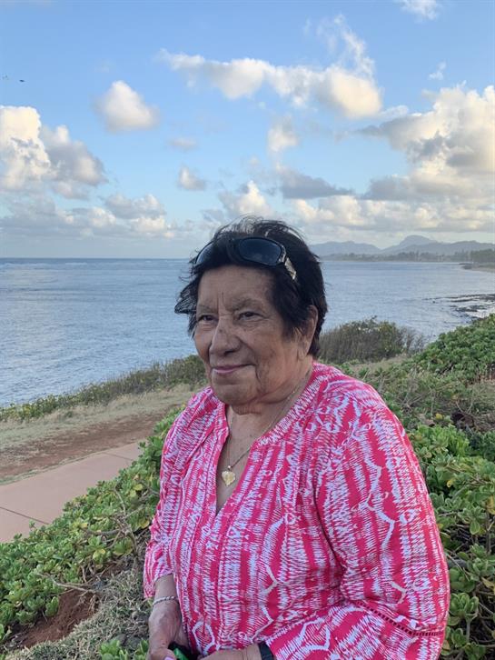 An elderly woman stands on a coastal path, smiling at the water under a clear sky.