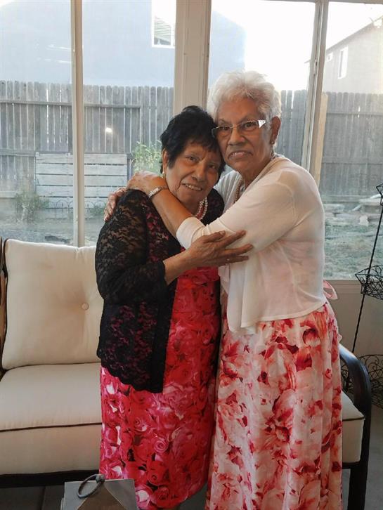 Two older women in floral dresses hug tightly, enjoying a warm moment in a bright living room.