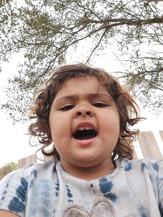 Young child plays joyfully outdoors, sitting under a tree and smiling widely, expressing happiness.