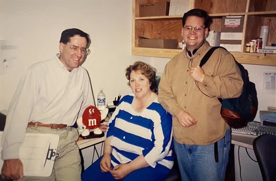 Three individuals pose in an office, showcasing a friendly atmosphere with colorful clothing.