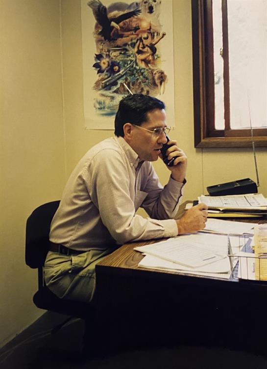 A man is on a phone call, taking notes at his cluttered desk in an office during daylight hours.